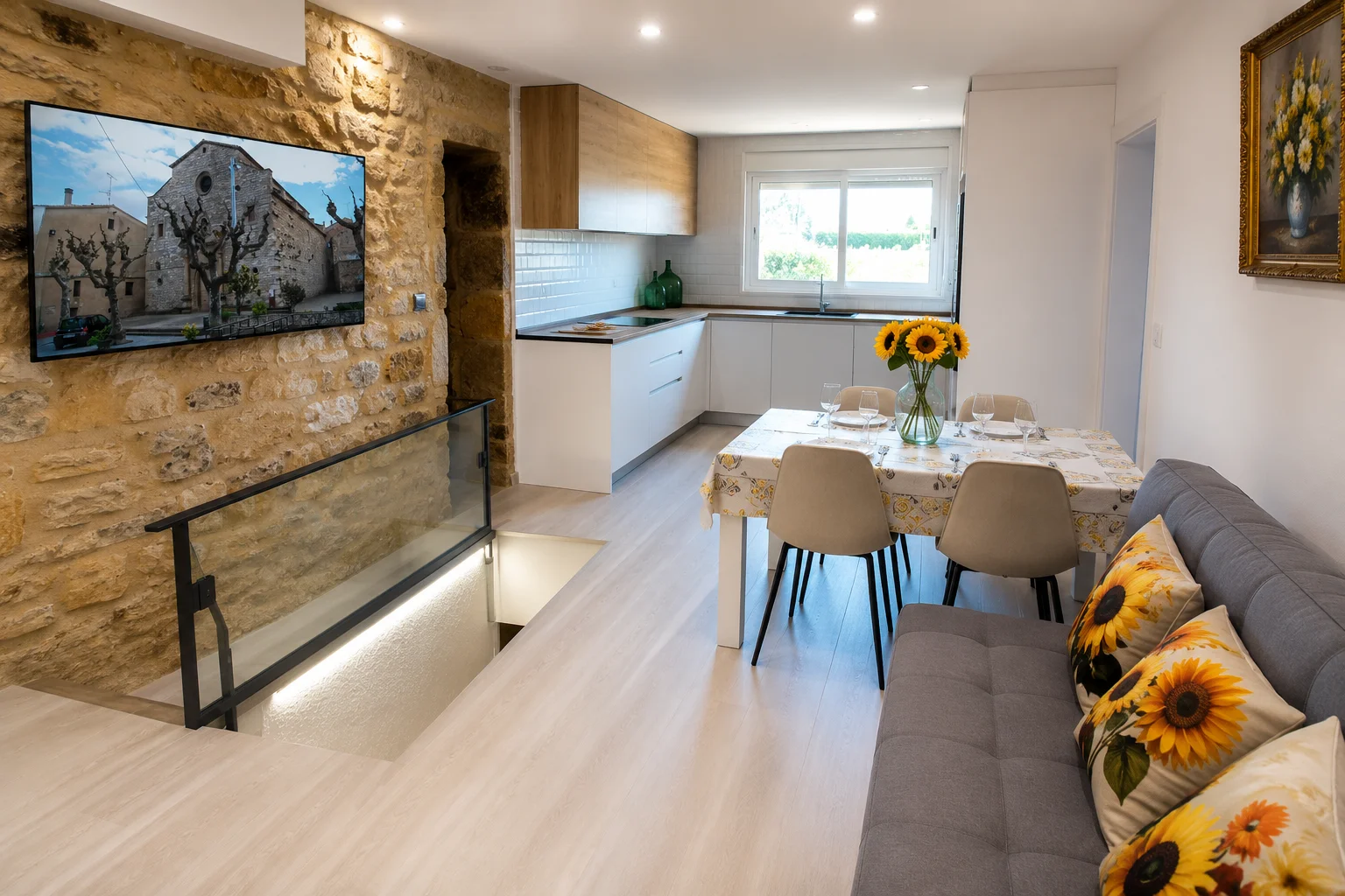 Salón comedor de Cal Casellas: pared de piedra con la televisión, la barandilla de cristal sobre la cisterna, la cocina blanca al fondo y la mesa puesta con girasoles